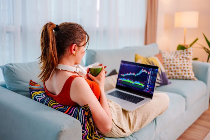 Young Businesswoman sits on the couch with her feet up, coffee cup in hand, checking the Bitcoin or exchange price chart on a digital exchange on her cell phone while doing research on a laptop monitor