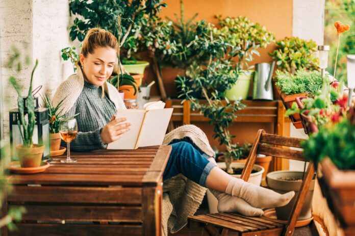 Young beautiful woman relaxing on cozy balcony, reading a book, wearing warm knitted pullover, glass of wine on wooden table